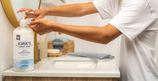 A person in a white shirt pumps liquid soap from a Kirks bottle on a bathroom counter, with a sink, mirror, and basket visible in the background.