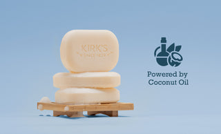 Three beige soap bars, one standing upright with Kirks Since 1839 stamped on it, are stacked on a small wooden soap dish. To the right, there’s an icon of coconut oil with the text Powered by Coconut Oil.