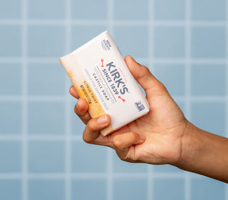 A hand holds a bar of Kirk’s Original Fresh Scent soap against a light blue tiled background. The soap is partially wrapped in white and yellow paper packaging.