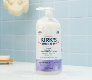 A bottle of Kirks 3-in-1 Head to Toe Nourishing Cleanser with lavender and vanilla sits on a bathroom counter, surrounded by soap suds, in front of a light blue tiled wall.