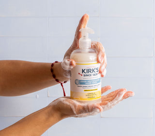 A pair of hands covered in soap suds holds a bottle of Kirks Odor Neutralizing Hand Wash with a pump dispenser against a light blue tiled background.