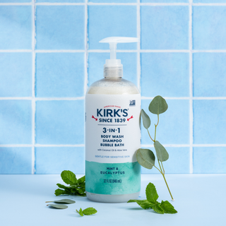 Mint & Eucalyptus - 3-in-1 Cleanser, featuring coconut oil, works as a body wash, shampoo, and bubble bath. Displayed in a clear pump bottle with fresh mint leaves and eucalyptus branches on a light blue surface before a blue tiled wall, the scene emphasizes its invigorating mint & eucalyptus scent.