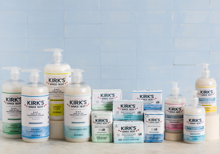 Assorted Kirk’s brand soaps and body washes are arranged on a counter against a light blue tiled wall; products include pump bottles and rectangular bar soaps with various labels and scents.