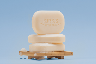 Three beige bars of soap with KIRKS SINCE 1839 embossed on the top bar are stacked on a wooden soap dish with some bubbles, set against a light blue background.