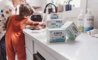 A child and adult wash hands at a kitchen sink while several bars of Kirk’s soap are stacked on the white countertop in the foreground. Bottles of liquid soap sit by the window in the background.