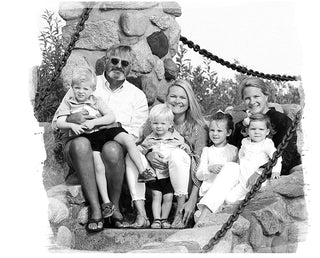 A black-and-white photo of a group of six people, including three adults and three young children, sitting and kneeling on stone steps outdoors, smiling at the camera. Shrubbery and chains are in the background.