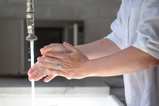 A person wearing a white shirt washes their hands with soap and water under a running faucet in a bright kitchen sink.